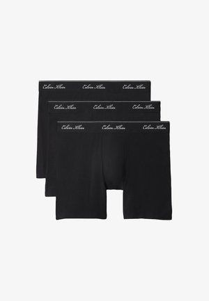 Hemdloser Mann mit lockigem Haar, der schwarze Boxershorts trägt, barfuß vor einem schlichten grauen Hintergrund steht.