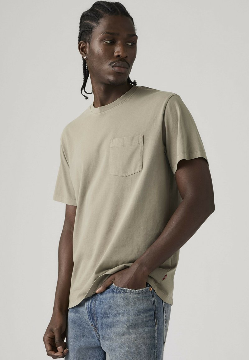 Jeune homme aux cheveux tressés portant un t-shirt beige avec poches et un jean bleu clair, debout avec une main dans la poche devant un fond uni.