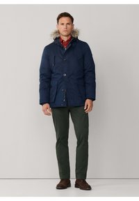 Parka azul marino con capucha forrada de piel, con cierre de doble botón, dos bolsillos delanteros y corte recto. Combinado con pantalones verdes y zapatos marrones.