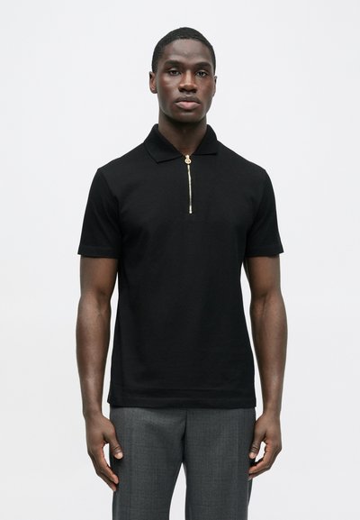 Juodas polo marškinėlis su trumpu užtrauktuku ir auksinės spalvos detalėmis. Pagamintas iš lygaus audinio, turi klasikinį apykaklę ir trumpas rankoves.