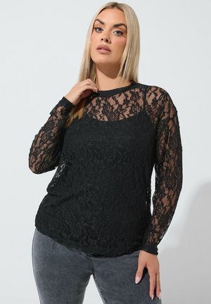 Blonde Frau trägt ein schwarzes langärmeliges Spitzenoberteil mit Blumenmuster über einem schwarzen Camisole, kombiniert mit grauen Jeans, posiert mit der Hand nahe am Gesicht.