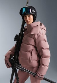 Roza pufer jakna s visokim ovratnikom i džepovima na patentni zatvarač, nošena s crnom kacigom i naočalama za skijanje, držeći skijaške štapove na sivom pozadini.