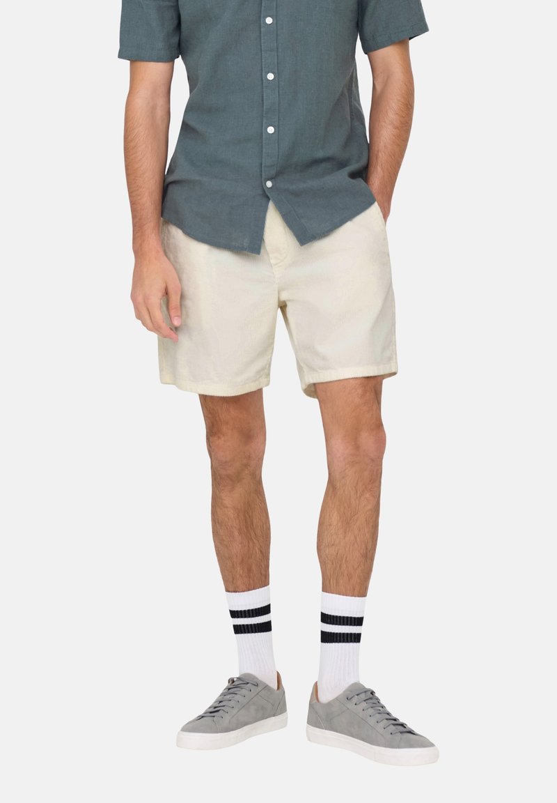 Shorts en coton beige avec des poches latérales, associés à une chemise bleue à manches courtes, des baskets grises et des chaussettes blanches avec des rayures noires.