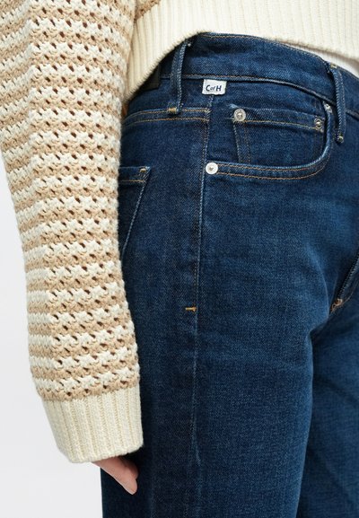 Tmavomodré riflové nohavice so žlto sfarbeným prešitím, dizajnom s piatimi vreckami a zapínaním na gombíky. Kombinované s béžovým textúrovaným pleteným svetrom.