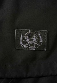 Černá látka s texturovanou koženou záplatou, na které je vyvýšený detailní design lebky s rohy, lemovaný prošitím.