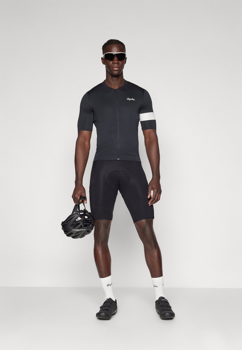 Jersey de ciclismo negro con cremallera corta, pantalones cortos a juego, acentos blancos y logo. El sujeto sostiene un casco negro, lleva gafas de sol y zapatos de ciclismo.