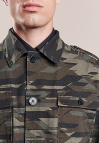 Mand iklædt en jakke med camouflage-mønster over en sort skjorte, der viser krave, bryst og knapdetaljer.