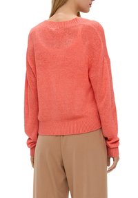 Koralowy sweter z dzianiny o luźnym kroju, z prążkowanymi mankietami i krawędzią oraz miękką fakturą. Noszony z beżowymi spodniami, prezentując swobodną sylwetkę.