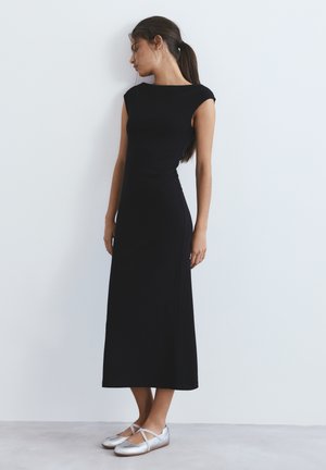 Nainen istuvassa mustassa hihattomassa mekossa ja hopeisissa balleriinoissa seisomassa valkoista seinää vasten, katse sivulle, hiukset poninhännällä.