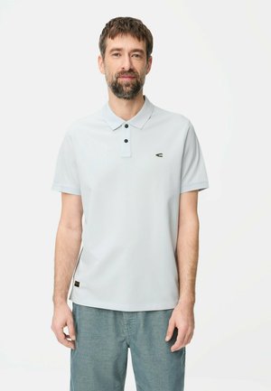 Mężczyzna z brodą, ubrany w jasnoszarą koszulkę polo z krótkim rękawem oraz niebieskie spodnie, stojący na jednolitym białym tle.