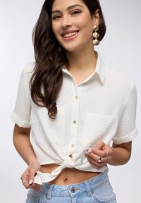 Camicia bianca, a maniche corte e texture, annodata in vita, abbinata a pantaloni corti in denim a vita alta. Due tasche e bottoni di perla.