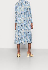 Blommönstrad långärmad klänning i ljusblått med gröna och krämfärgade accenter. Längd nedanför knäna, matchad med ljusgula klackar.