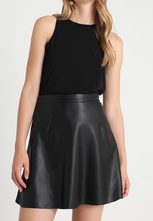 Femme portant un haut noir sans manches rentré dans une jupe en cuir noire taille haute en forme trapèze sur un fond uni.