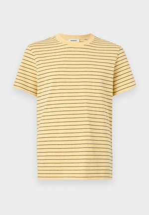 Homme aux cheveux bouclés portant un t-shirt jaune à rayures, un pantalon large noir et des baskets blanches, debout les mains dans les poches en regardant vers le bas.