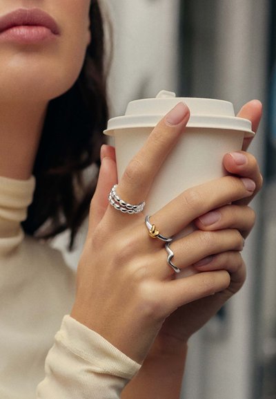 Γυναίκα που φοράει ασημένια δαχτυλίδια με χρυσές λεπτομέρειες κρατάει ένα λευκό χάρτινο κυπελλάκι καφέ με καπάκι κοντά στο πρόσωπό της.