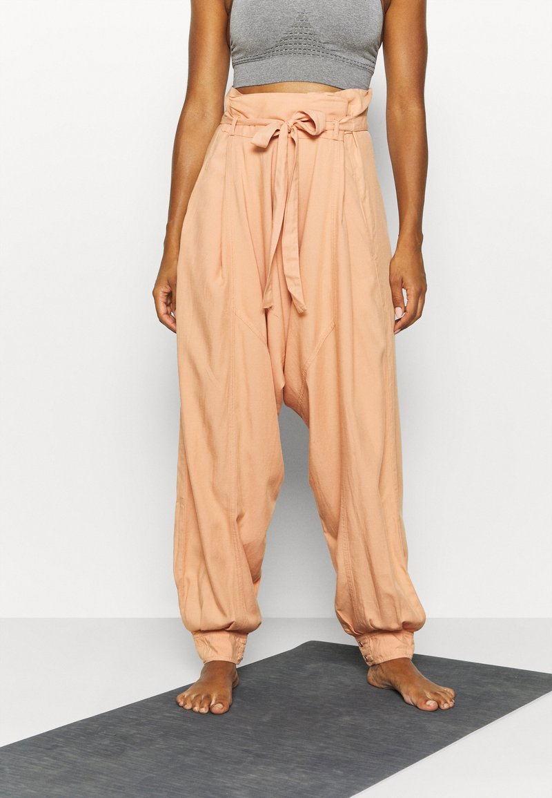 Femme debout pieds nus sur un tapis, portant un pantalon ample couleur pêche avec une taille nouée et un top de sport gris court.