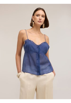 Femme portant un haut sans manches bleu marine transparent avec boutons sur le devant et bordure en dentelle, ainsi qu'un pantalon large crème, posant les mains dans les poches.