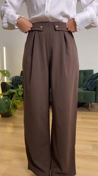 Άτομο που φοράει καφέ παντελόνι με ευρύ πόδι και ψηλή μέση με χρυσά κουμπιά, τα χέρια του βυθισμένα στη μέση, λευκό πουκάμισο, εσωτερικός χώρος με φυτά και καναπέ.