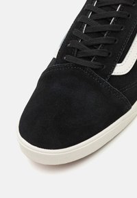 Baskets en daim noir avec accents en cuir blanc, présentant un bout rond et des lacets noirs, ainsi qu'une semelle en caoutchouc texturée légèrement surélevée.