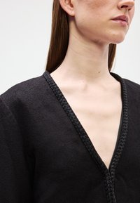 Mulher vestindo uma peça preta texturizada em V, com acabamento trançado, mostrando o pescoço, a linha da mandíbula e a clavícula contra um fundo liso.