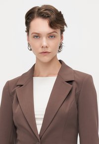 Hnědý upravený blazer s ostrými klopami, zapínáním na jeden knoflík a přiléhavým střihem, kombinovaný s bílým topem s vysokým límcem pod ním.