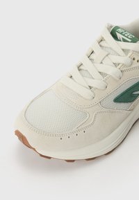 Light beige sneaker with mesh and suede materials, green accents, round toe, flat laces, and a textured brown rubber sole.