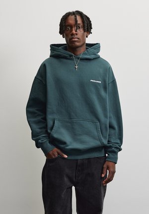 Junger Mann mit geflochtenem Haar, der einen dunkelgrünen Hoodie mit Fronttasche und schwarze Hose trägt, vor einem schlichten hellen Hintergrund stehend.