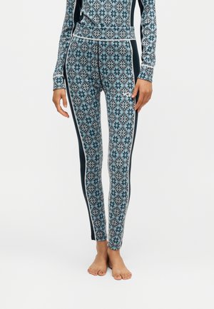 Kvinde iført blå og sorte mønstrede leggings med geometrisk blomstermønster og sorte sidepaneler, stående barfodet på en hvid baggrund.