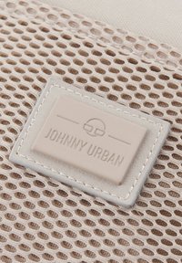 Beige mesh-tyg med en slät, ljusgrå etikett präglad med "JOHNNY URBAN." Har sydda kanter och en texturerad bakgrund.