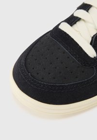 Fekete velúr sneaker, perforált orral, krémszínű fűzőkkel és kontrasztos, törtfehér talppal. Görbült dizájnt és varrott részleteket mutat.