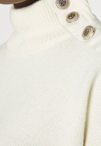 Krämfärgad polokrage tröja med ribbad struktur och dekorativa guldknappar på axeln, som framhäver en strukturerad men ändå mjuk design.