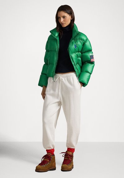 Zelena puhaste bunda s črtastim vzorcem, črn rolka, kremne trenirke in rjavi pohodniški čevlji z rdečimi vezalkami in poudarki.