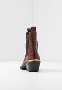 Bottine à talon bas en cuir synthétique marron avec motif crocodile, dotée de panneaux élastiques sur les côtés et d'un accent métallique sur le talon, sur fond blanc.