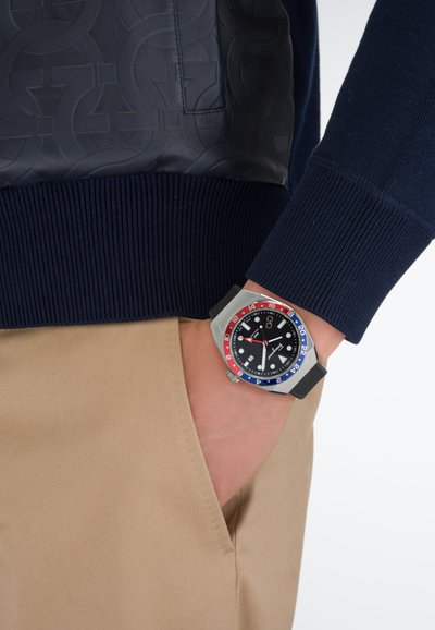 Hõbedane sportlik kell musta sihverplaadi, punase ja sinise raamikujundusega, kantud käel. Hall kummirihm, taustal tumesinine kampsun ja beežid püksid.