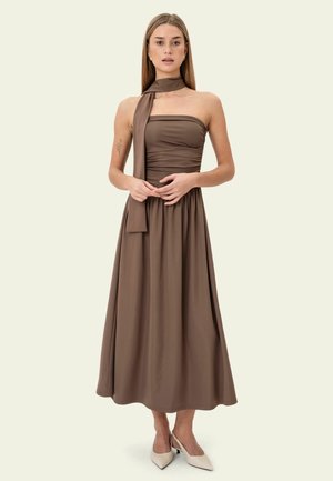 Kvinde med langt, glat hår iført en brun, skulderfri midi-kjole med tilhørende tørklædedetalje og beige spidse hæle, stående mod en ensfarvet baggrund.