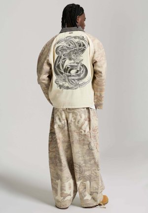 Persona con el cabello trenzado lleva pantalones anchos de camuflaje beige con texto blanco y una chaqueta crema que presenta un detallado diseño de serpiente negra en la espalda.