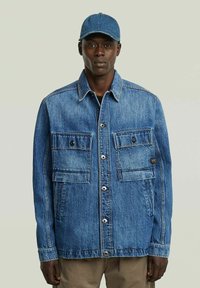 Un uomo che indossa una giacca di jeans blu con grandi tasche frontali e un cappellino di jeans blu abbinato, in piedi su uno sfondo neutro.