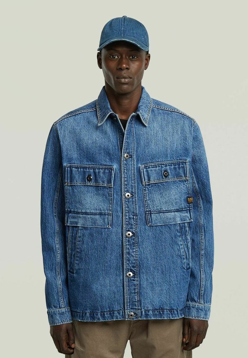 Un uomo che indossa una giacca di jeans blu con grandi tasche frontali e un cappellino di jeans blu abbinato, in piedi su uno sfondo neutro.