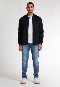 Zwarte bomberjack met ritssluiting, geribbelde manchetten, gedragen over een wit T-shirt, gecombineerd met lichtblauwe jeans en witte sneakers.