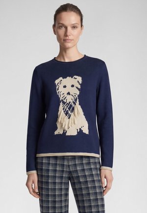 Frau trägt einen marineblauen Pullover mit einer beigen Hunde-Grafik und Fransen, kombiniert mit blau-weiß karierter Hose vor schlichten grauem Hintergrund.