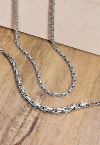 Collane in metallo argentato con link rettangolari, caratterizzate da una superficie liscia e una finitura lucida, disposte su una superficie di legno.