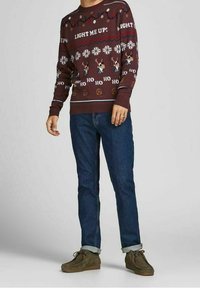 Pull bordeaux avec un imprimé festif incluant des flocons de neige et des rennes, portant le texte "LIGHT ME UP!" et des poignets côtelés. Porté avec un jean foncé et des chaussures marron.