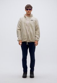 Φούτερ μπεζ ανοιχτού χρώματος με φερμουάρ μπροστά, με τσέπη στο αριστερό στήθος και ελαστικά μανίκια, συνδυασμένο με σκούρο μπλε παντελόνι και μαύρες μπότες.