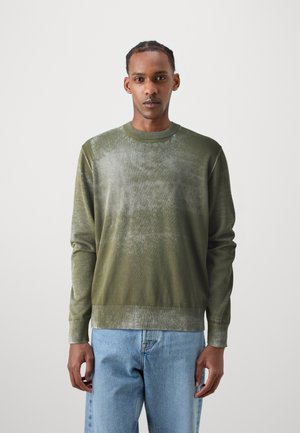Olive green knitted sweater featuring a gradient texture and ribbed cuffs, paired with light blue denim jeans.