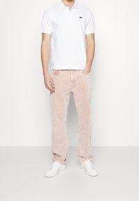 Homme portant un polo blanc, un pantalon en velours côtelé rose clair et des baskets blanches, se tenant avec les mains dans les poches devant un fond uni.