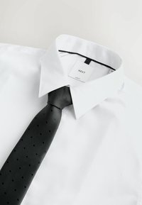 Chemise blanche slim fit avec col large et cravate noire présentant un motif discret à pois, posée à plat sur un fond clair.
