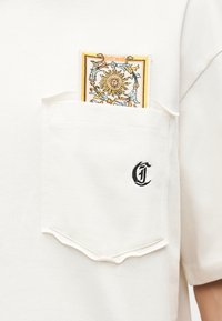 Λευκή τσέπη πουκάμισου με μαύρο κεντητό γράμμα "C" και διακοσμητική επιφάνεια με σχέδιο ήλιου και λουλουδιών που προεξέχει.