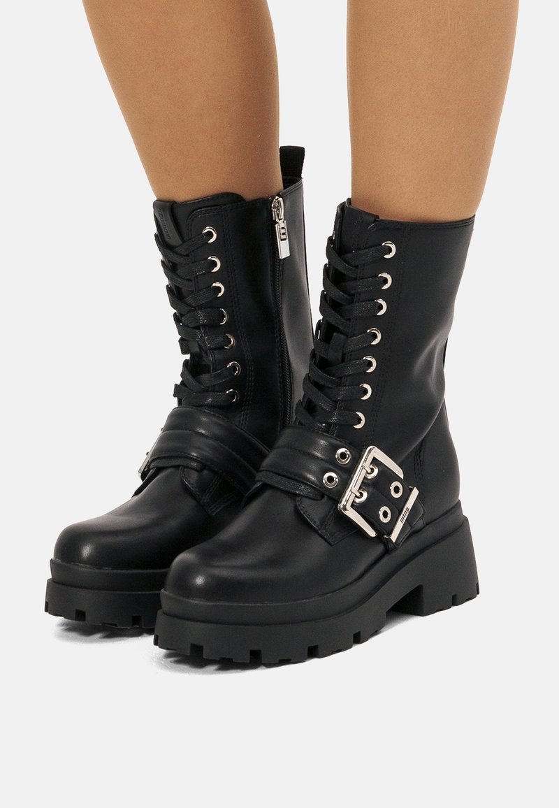 Bottes noires à lacets montant à la cheville, avec semelles épaisses à crampons, fermetures éclair latérales et larges brides à boucles argentées portées sur des jambes nues.