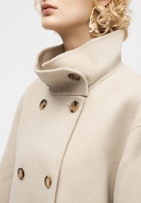 Γυναίκα με σγουρά ξανθά μαλλιά φοράει μπεζ παλτό από μαλλί με ψηλό γιακά και μεγάλα κουμπιά σε σχέδιο χελώνας, συνδυασμένο με χρυσούς σκουλαρίκια σε σχήμα αφηρημένου κρίκου.