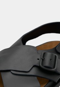 Μαύρο δερμάτινο σανδάλι με ευρύ σχεδιασμό λουριού, ρυθμιζόμενο αγκράφα και λείο υλικό με καφέ πάτο.